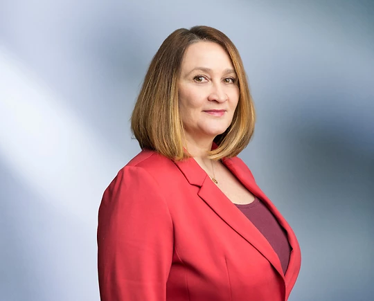 A woman, Inna Musser, in a red blazer and maroon top is looking at the camera, standing against a blurred blue and white background.