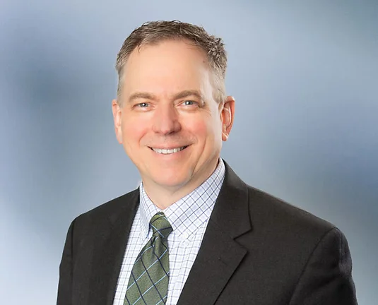 A middle-aged man, Thomas Huyck, smiles at camera, wearking a black sport coat with stripped patterned collared shirt and green necktie in front of blue gray blurred background