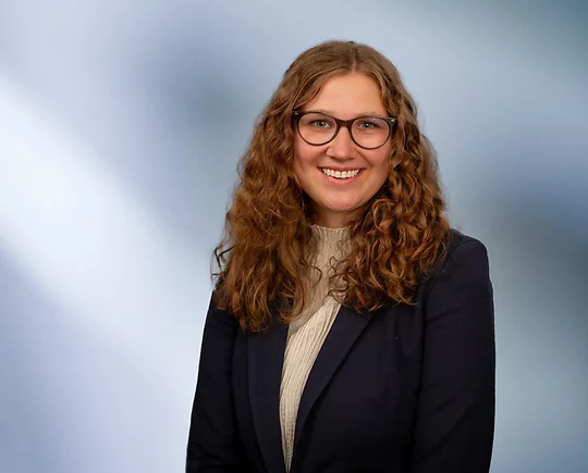 A woman, Mackenzie Almassian, with long, wavy blonde hair, wearing a navy blazer and white top, is smiling at the camera, standing against a blurred blue and white background.”
