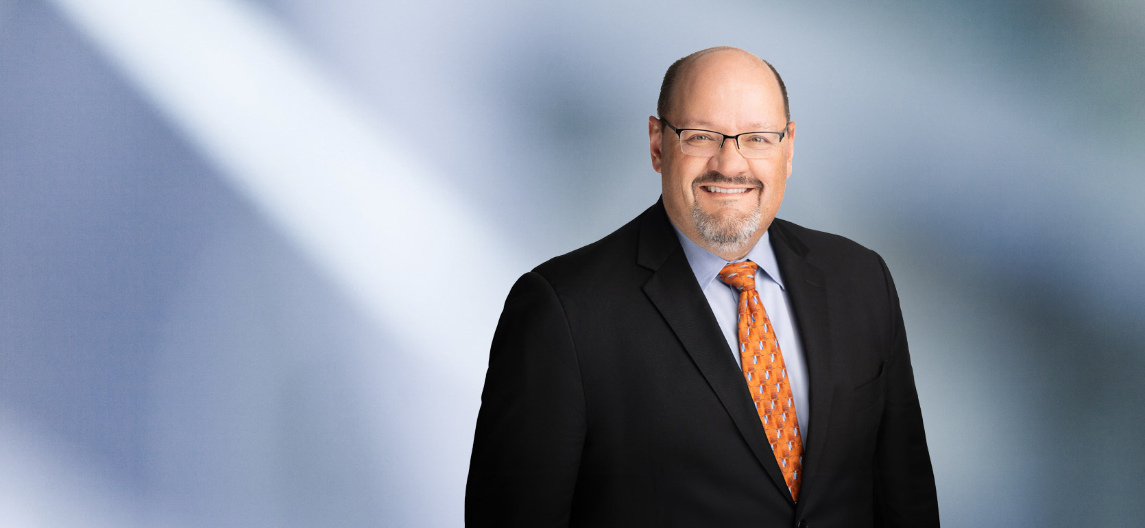 A man, Kevin Roragen, in a black suit, light blue shirt, and orange patterned tie is smiling at the camera, standing against a blurred blue and white background.