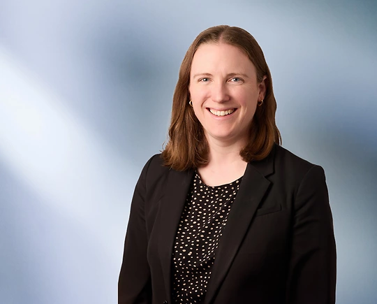 woman, Rachael Kuleima Klein, with brown hair, wearing a black blazer, smiling against a blurred blue and white background"