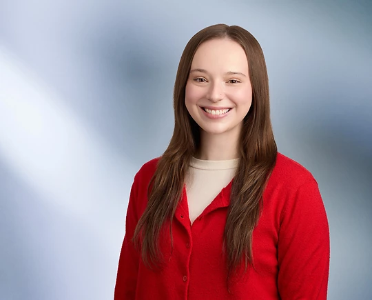 A young woman, Emma Benoit, in a red cardigan and cream top is smiling at the camera, standing against a blurred blue and white background.