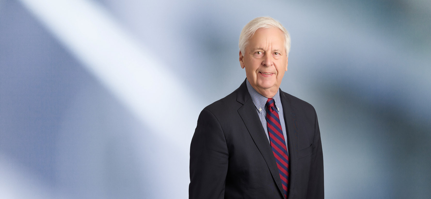 A man, Lynwood Vandenbosch, in a dark suit, checkered shirt, and red and blue striped tie is smiling at the camera, standing against a blurred blue and white background.