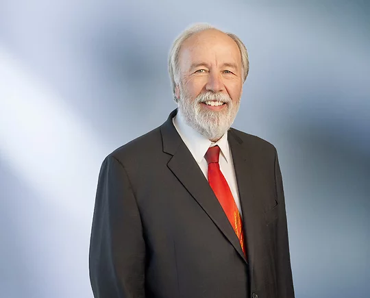 man, Dirk Beckwith, with a white beard wearing a dark suit, white shirt, and bright red tie, standing against a softly blurred blue and white background, smiling warmly at the camera.