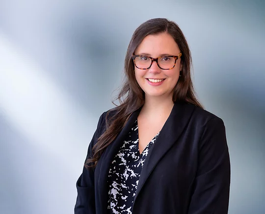 A brown-haired woman, Amy Marinkovski, wearing turtle frame glasses, black sport coat and black and white zigzag pattern shirt, smiles at camera in front of blue gray background