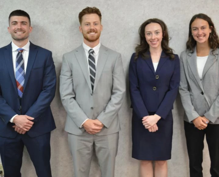 Four young professionals stand in a row against a light gray wall, smiling and dressed in formal business attire.