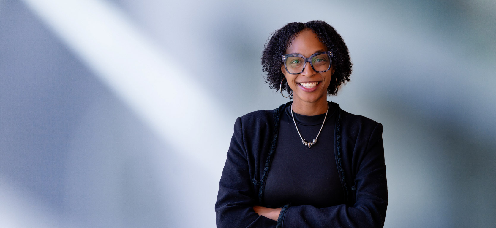 African American woman, Kanielle Pounds, with large black rimmed glasses smiles at camera in front of blue gray background wearing a black sweater and black shirt