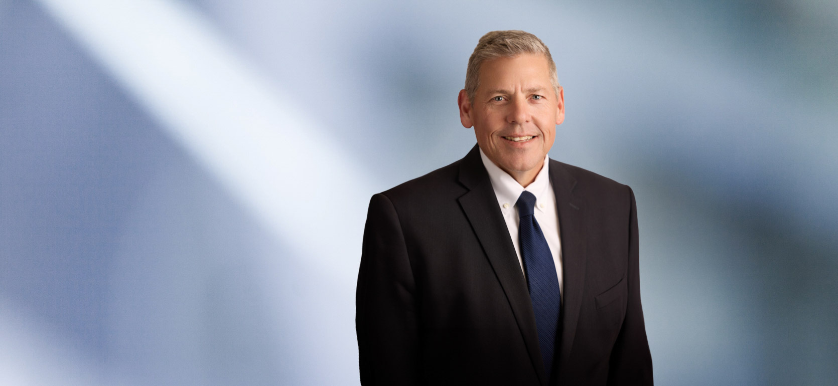 "A man, Michael Homier, with short, light brown hair, wearing a dark blazer, is smiling at the camera, standing against a blurred blue and white background."