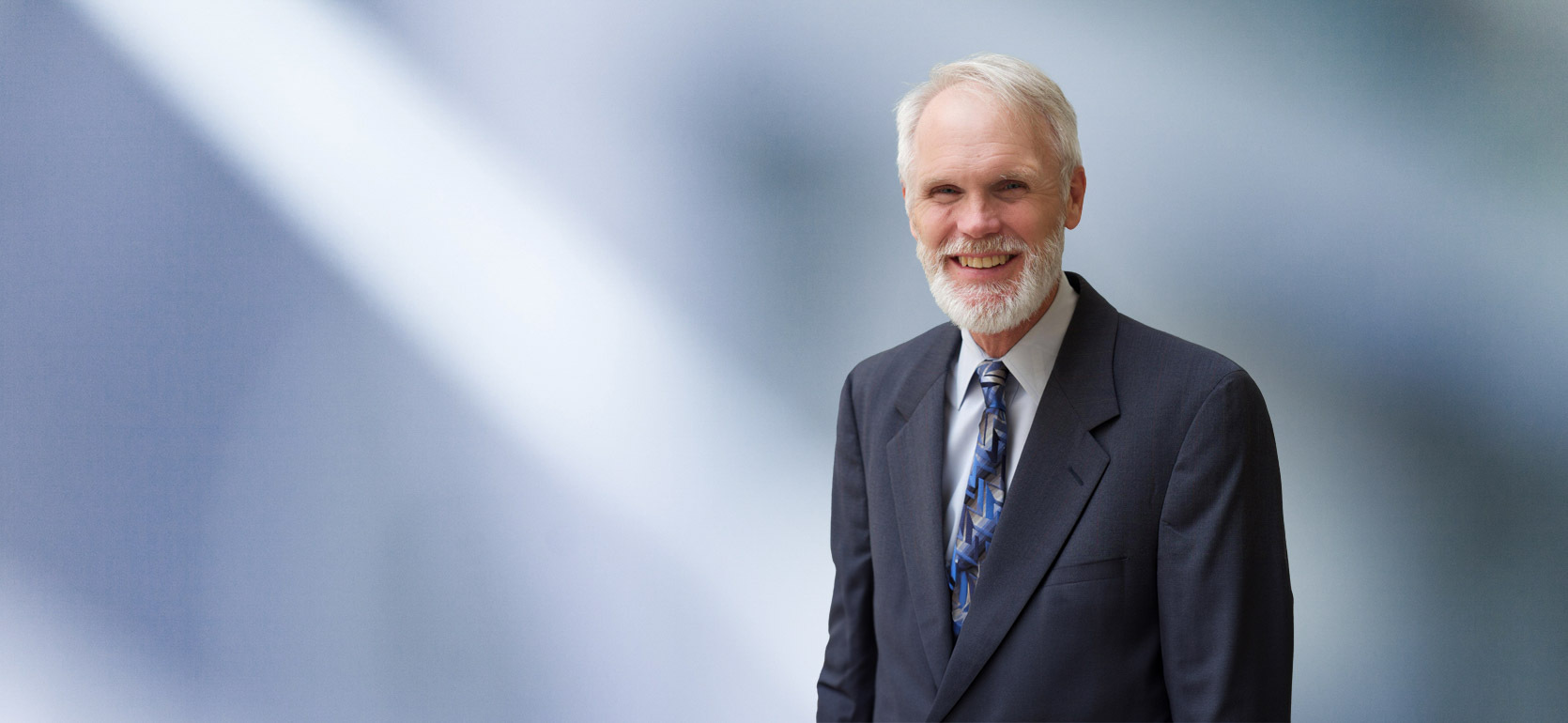 A man, Charles Barbieri, in a dark suit, white shirt, and patterned blue tie is smiling at the camera, standing against a blurred blue and white background.