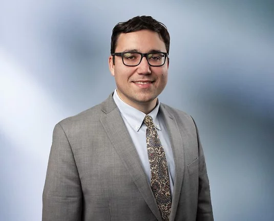 A professionally dressed man, Jeff Schultz, wearing glasses, a light gray suit, and a patterned tie, smiling against a softly blurred blue gradient background.
