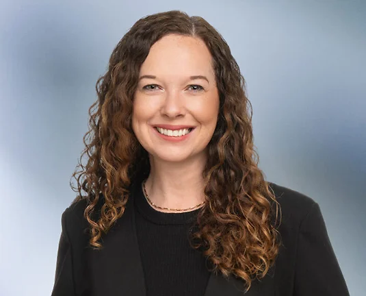 A woman, Laura Genovich, with curly brown hair, wearing a black blazer and black top, is smiling at the camera, standing against a blurred blue and white background.