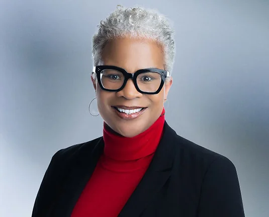 A woman, Dora Brantley, in a red turtleneck and black blazer is smiling at the camera, standing against a blurred blue and white background.